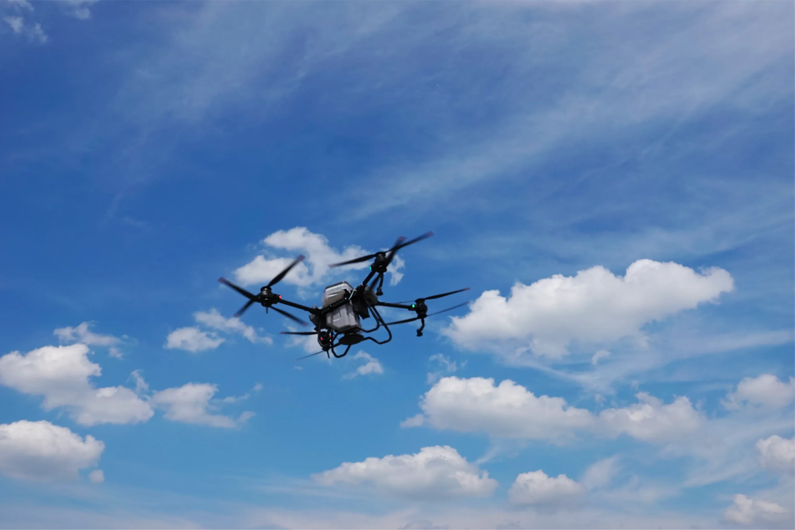 Drone agricole en plein vol dans un ciel bleu avec nuages, utilisé pour l’épandage de précision.